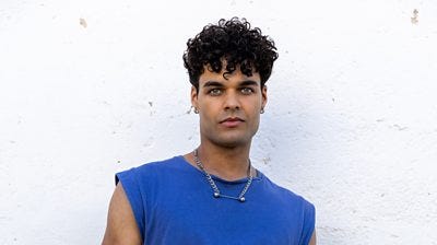 Ruben wears a blue tank top with a chunky silver chain. He has hoop earrings in both ears 
