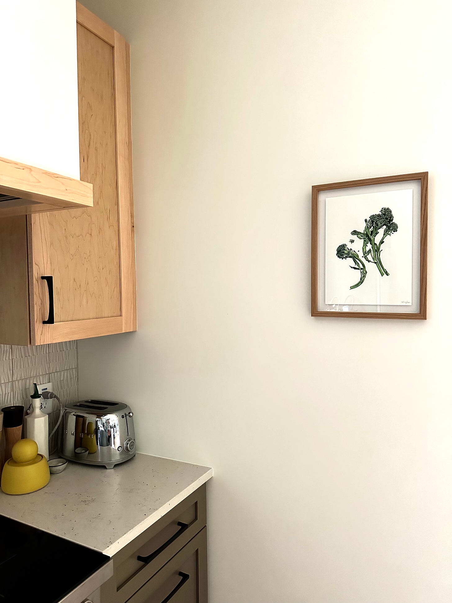 A stove and countertop with wooden cabinets is to the left, while on the wall to the right hangs a watercolor painting of broccolini, in a thin wooden frame. 