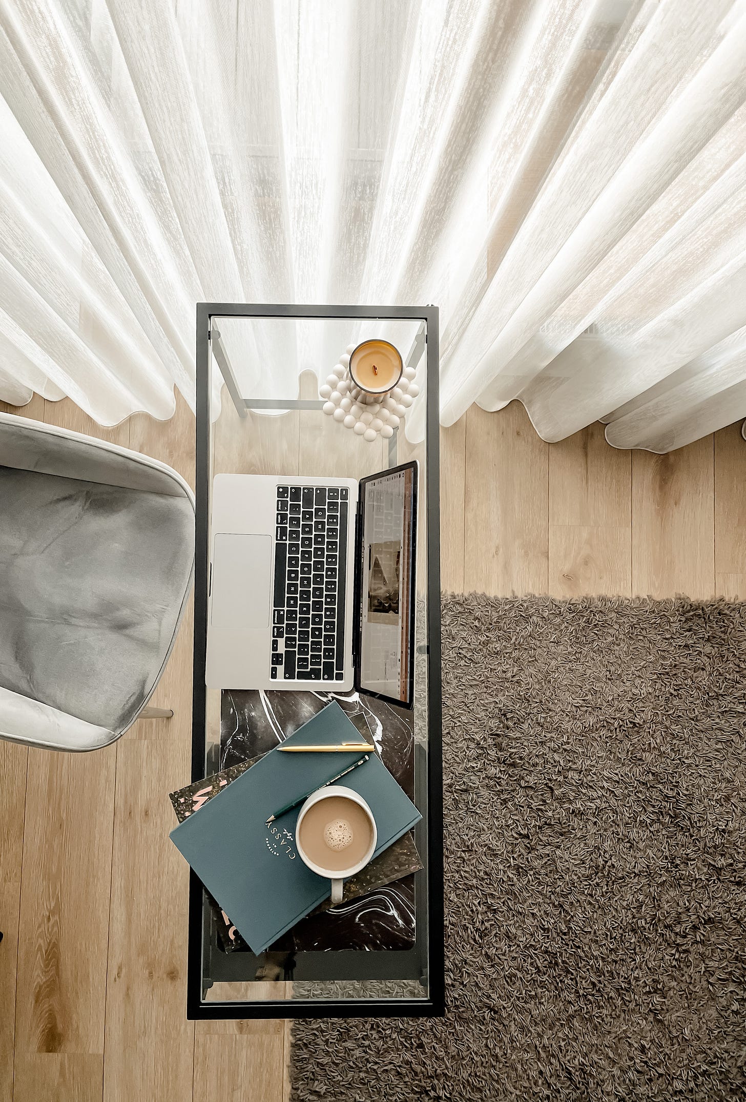 An overhead view of a glass desk in a home office with natural light coming through white linen curtains. A laptop sits on the desk with a cup of coffee and notebooks spread out. An overhead view of a glass desk in a home office with natural light coming through white linen curtains. A laptop sits on the desk with a cup of coffee and notebooks spread out.