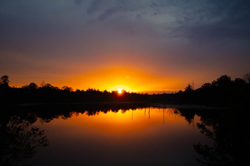 A sunset with a purple and orange and reddish orange sky over water that reflects the sky, with some dark shadowy trees along the far edge of the water