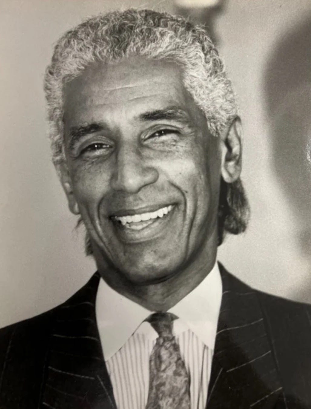 Black and white photo of Khadijah's father. He's smiling, wearing a dark suit with a striped shirt and paisley tie. His hair is wavy, and clearly salt and pepper.