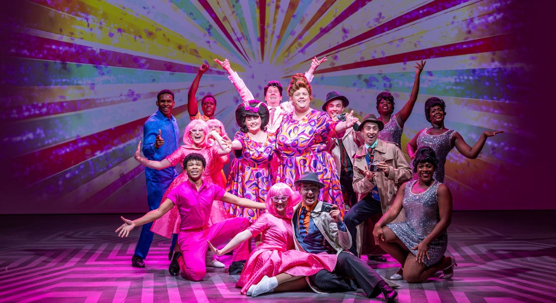 The cast of Hairspray wearing pink and blue outfits, posing in a crowd together on a stage with pink lighting The cast of Hairspray wearing pink and blue outfits, posing in a crowd together on a stage with pink lighting
