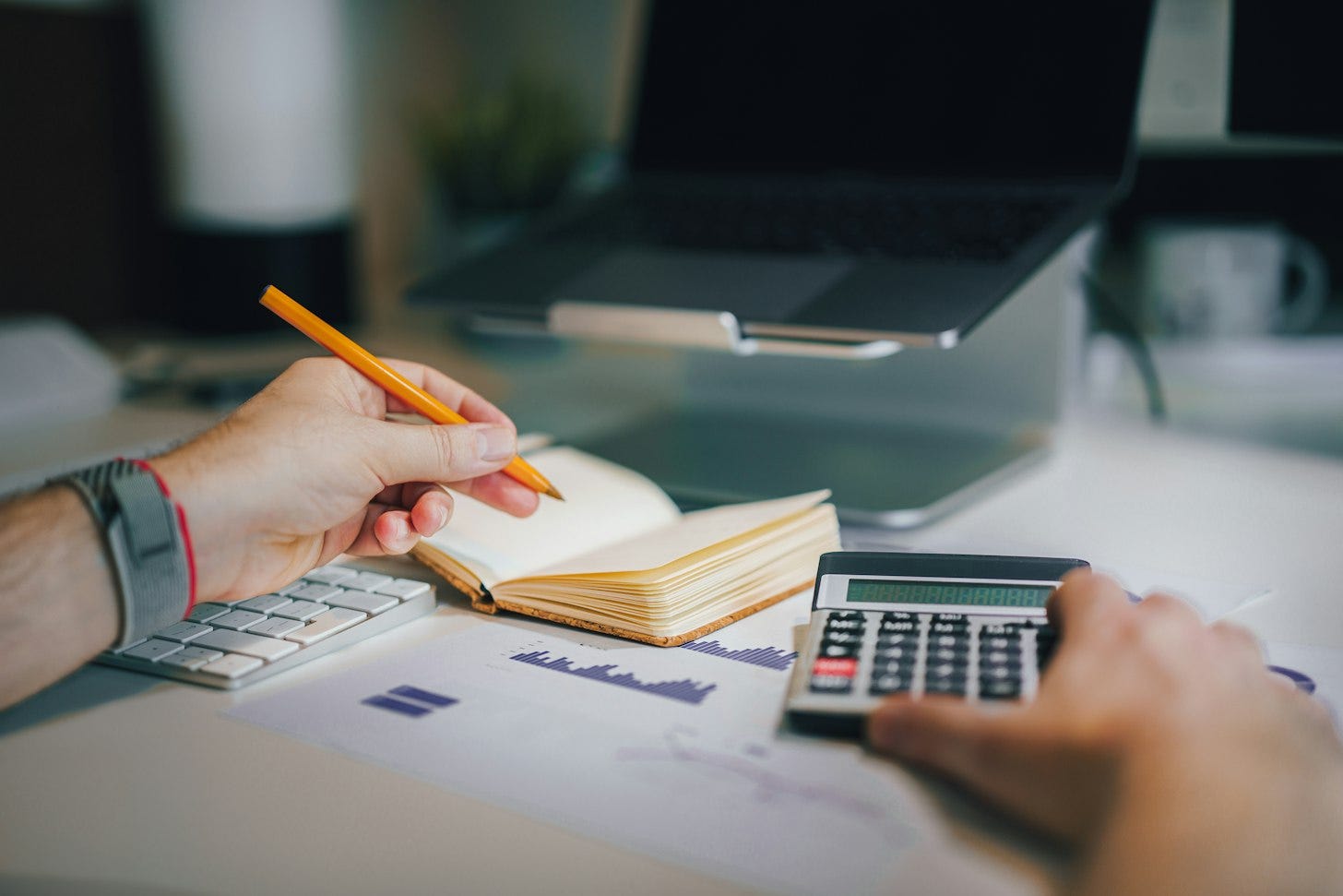 A person writes in a paper notebook with a pencil next to a calculator, with an open laptop in the background. A person writes in a paper notebook with a pencil next to a calculator, with an open laptop in the background.