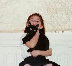 Lauren as a small child is smiling directly into the camera as she holds her small, black cat. Lauren is smiling.