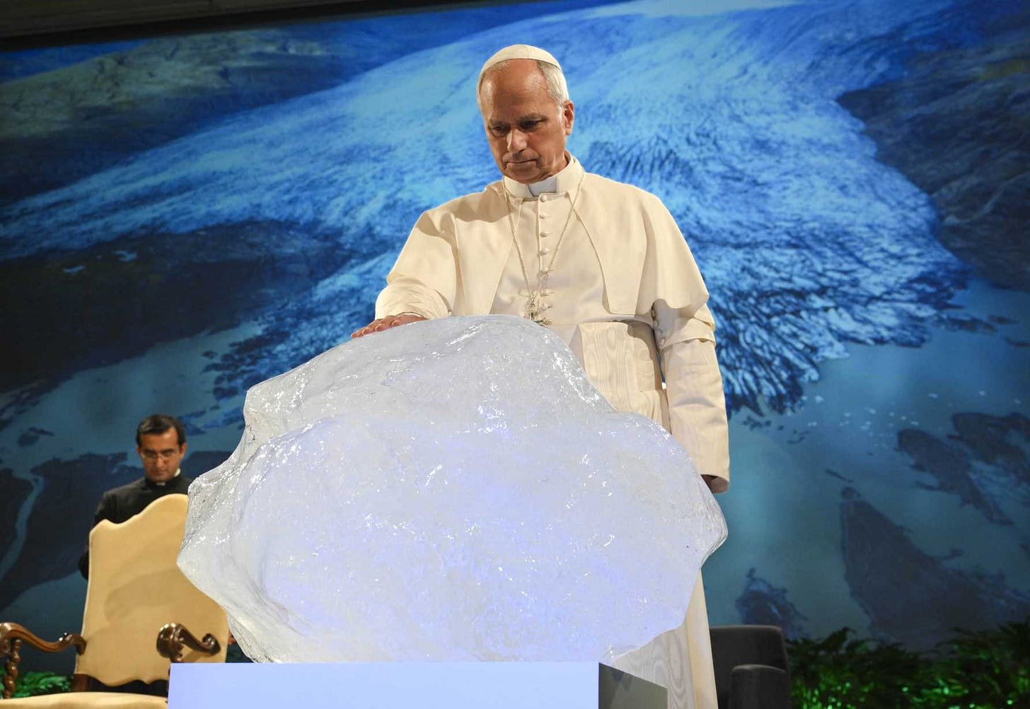 Pope Leo XIV, in white papal robes, places his right hand on a several-hundred-pound piece of ice on an onstage stand, giving the traditional blessing used for holy water. Projected on a wall-sized screen behind him is a photo of Earth as seen from space. Pope Leo XIV, in white papal robes, places his right hand on a several-hundred-pound piece of ice on an onstage stand, giving the traditional blessing used for holy water. Projected on a wall-sized screen behind him is a photo of Earth as seen from space.