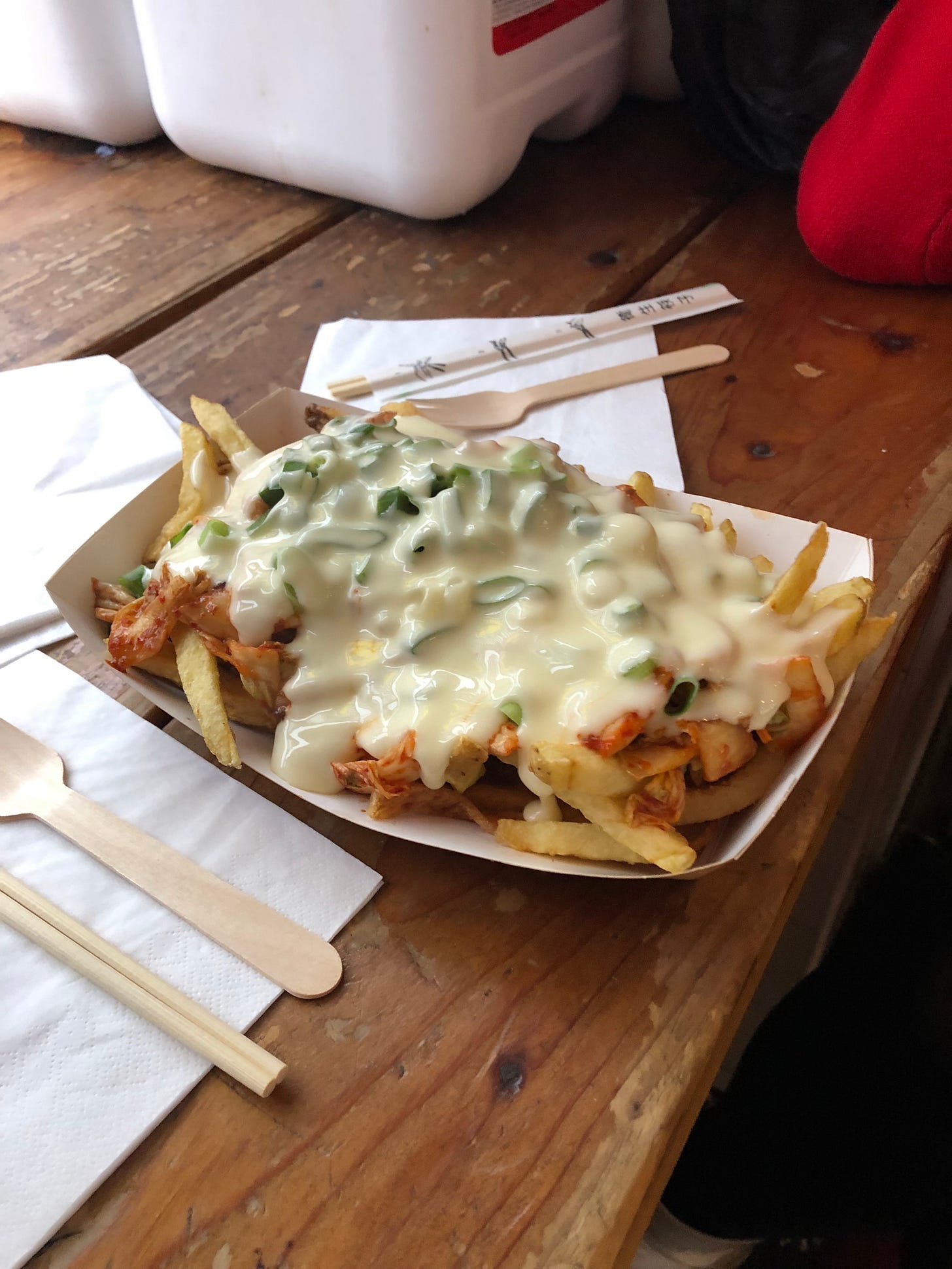 A paper dish crammed with thin chips, covered with kimchi and then smothered in a loose cheese and spring onion sauce. Wooden cutlery and chopsticks lay on the table beside. A red knit elbow is bent against the table in the top corner. 