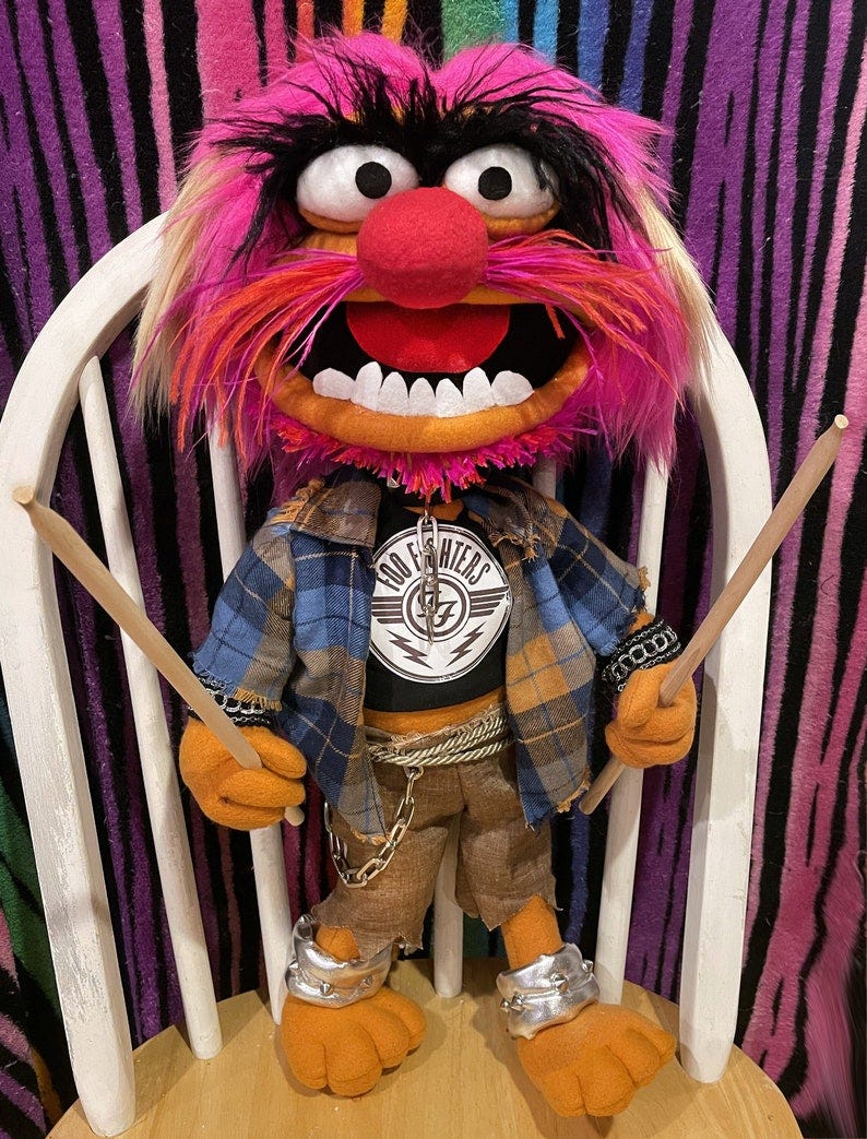 May include: A pink and orange puppet with black hair and a red nose. The puppet is wearing a blue and brown plaid shirt, a black shirt with a silver chain and a patch that says 'Foo Fighters', brown pants, and silver bracelets. The puppet is holding two drumsticks.