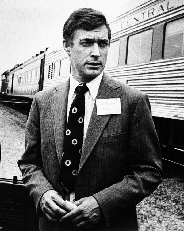 A black-and-white photo of a man in a suit and tie, with a name tag attached to his left lapel, standing in front of rail cars. A black-and-white photo of a man in a suit and tie, with a name tag attached to his left lapel, standing in front of rail cars.