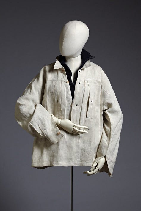 1) Classic black-and-white portrait of Gluck in a loose white shirt/smock, looking down and to the side. Their expression is thoughtful, with shadows adding depth. They have short dark hair, and they are holding a cigarette in their right hand. There is a dark shirt sticking out from under the larger shirt, with both collars sticking up. 2) A white mannequin displays a vintage, cream-colored linen shirt/smock with a black collar and buttoned cuffs, evoking a classic, timeless style. It's positioned in the same style Gluck was wearing it in the previous photo, including the collars sticking up. The mannequin head is also looking to the side, like Gluck was. 3) A pair of vintage brown leather shoes with buckles and low heels on a neutral background, conveying a sense of timeless style and craftsmanship. 4) Five vintage dresses/gowns hang on a black clothing rack with a small jacket. Patterns include floral, paisley, and abstract designs, evoking a nostalgic and elegant feel. 5) A white mannequin displays an elegant black evening gown with a lace overlay. The dress features long sleeves and a floor-length skirt, conveying sophistication.