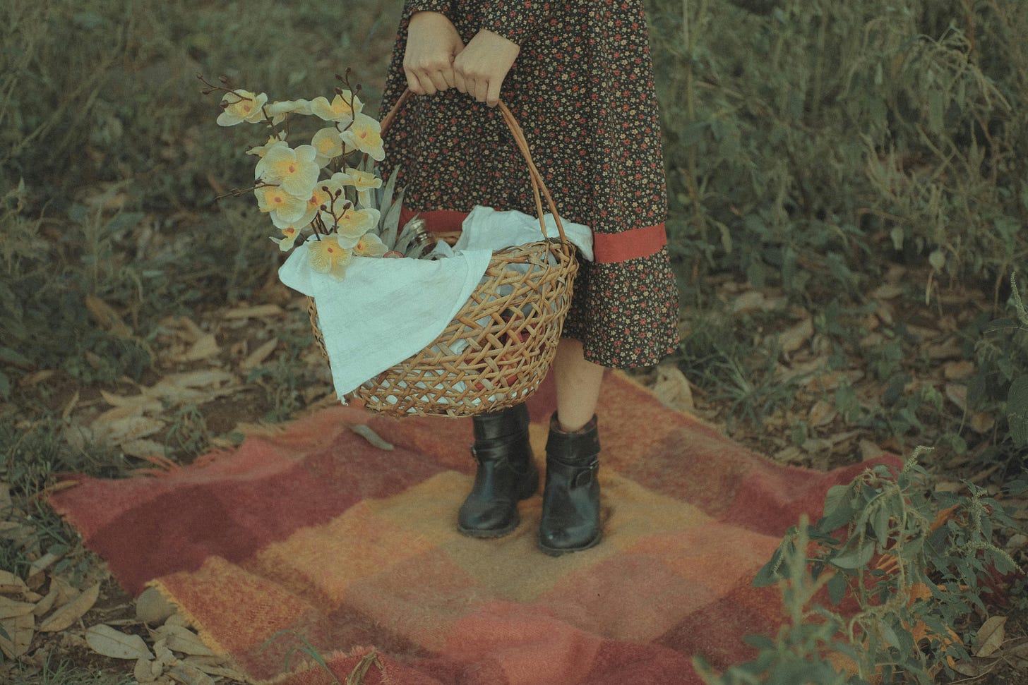 Shown from the hips down, a woman in a long floral dress stands on a plaid blanket in the grass. With both hands, she holds a wicker basket that has yellow orchids in it. 