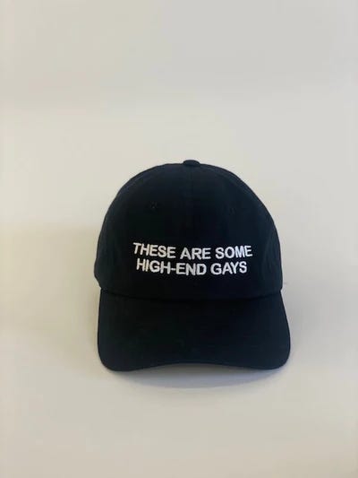 Two black hats with white embroidery. One says, "These are some high-end gays" and one says, "RIP Tanya McQuoid".