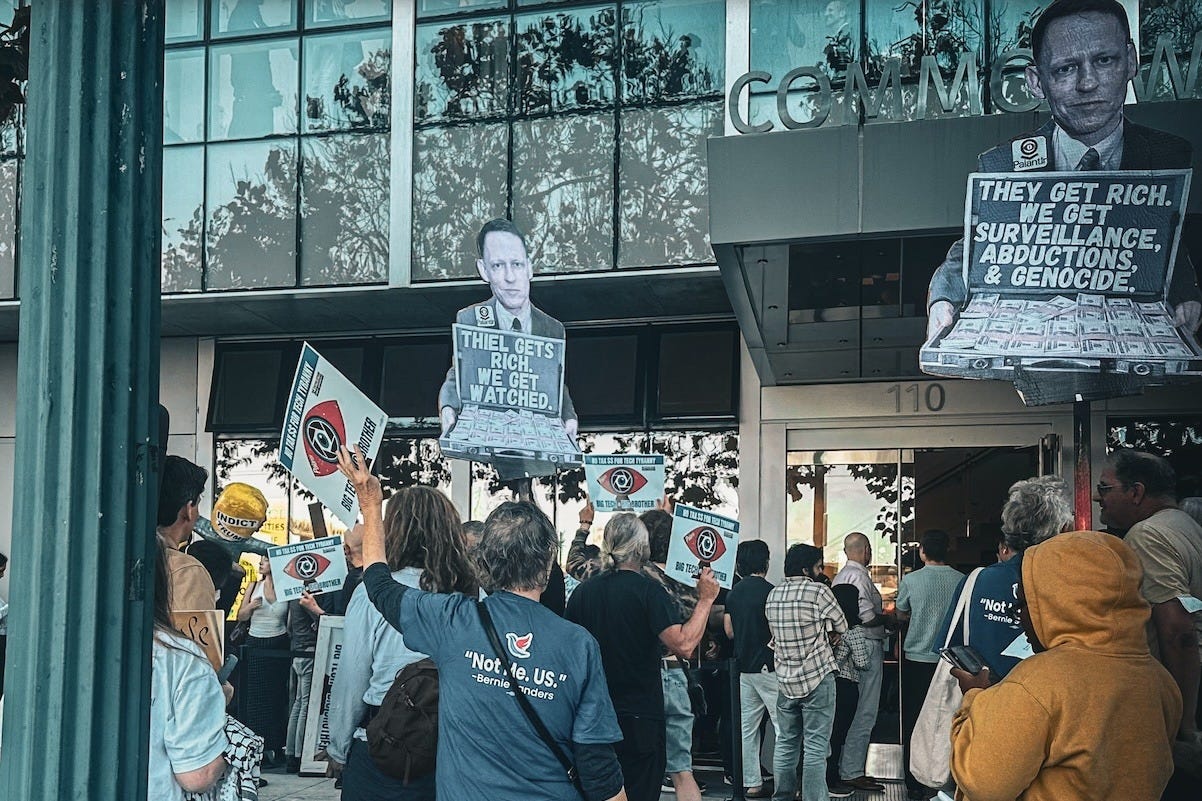 Protesters outside San Francisco’s Commonwealth Club hold signs featuring Peter Thiel and criticizing Palantir’s role in surveillance, immigration enforcement, and military operations.