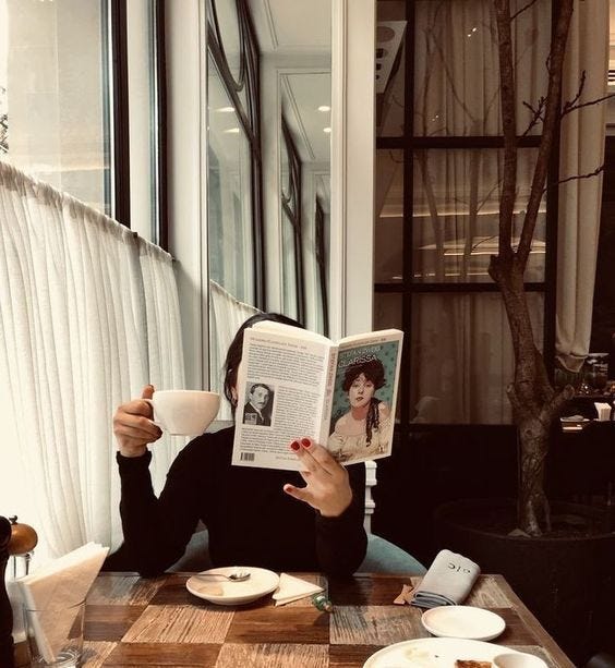 This may contain: a woman sitting at a table reading a book and holding a coffee cup in front of her face