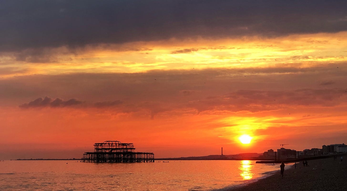 Silhouette of Brighton's burnt West Pier against dramatic sunset with dark storm clouds above, golden light reflecting on calm water, and a solitary figure on the beach in the foreground