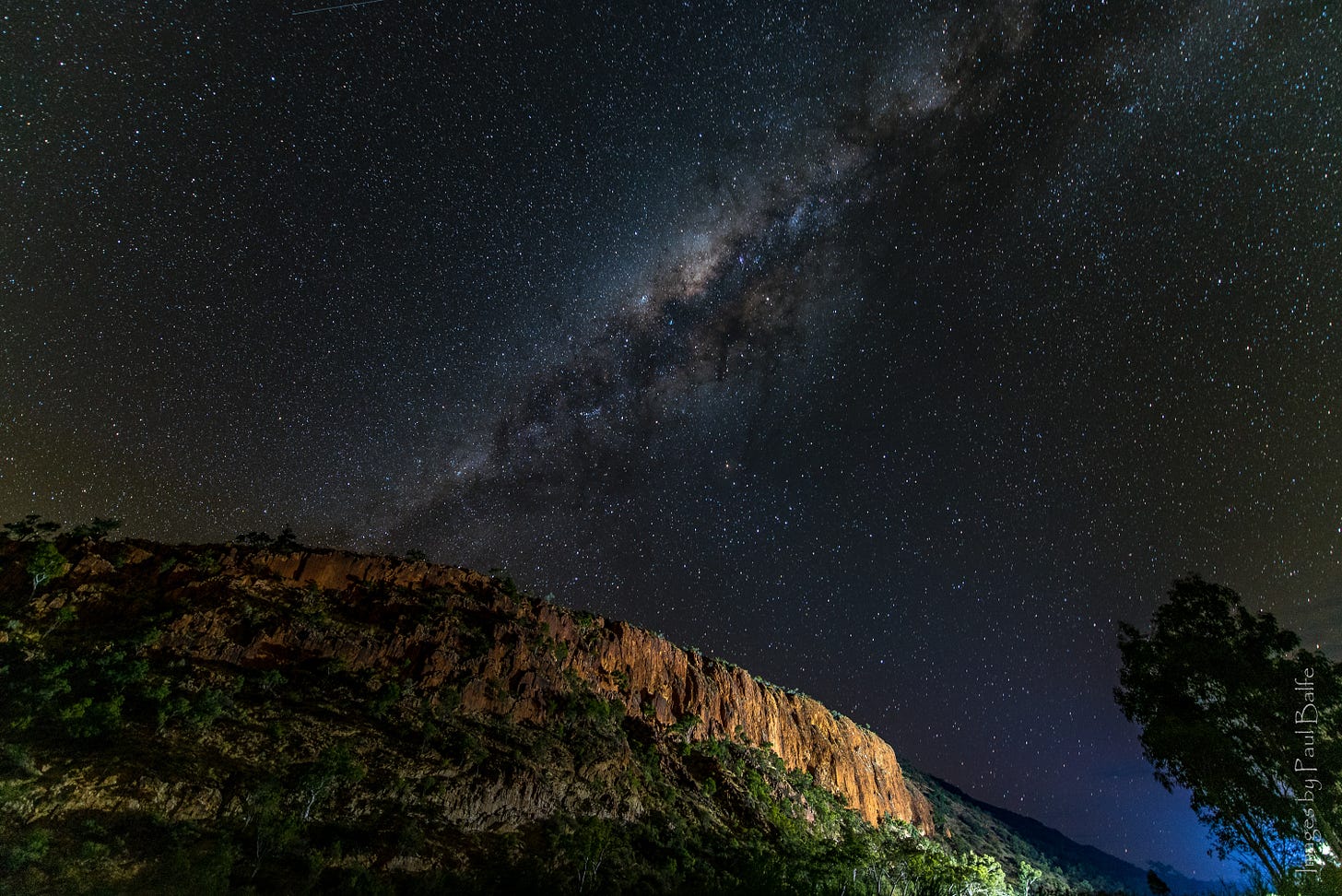 A photograph of a landscape at night time. The foreground has a dimly lit mountain ridge with greenery along the bottom. The sky is a deep, dark blue and features hundreds of small stars, with the shading of the Milky Way visible.