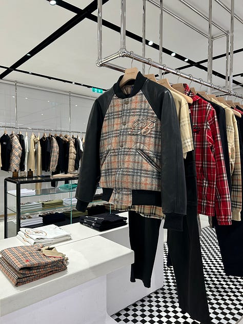 Three-photo gallery from an Intern Pierre primary channel-check store visit to a Burberry flagship, documenting the gap between the company's articulated heritage refocus narrative and the actual visual merchandising on the floor. Left photo: the women's handbag wall positioned at the storefront entry zone, with structured leather Burberry shoulder bags, hobo bags, and check-pattern canvas backpacks displayed on backlit white wall shelves above a glass jewelry display case and a beige suede storage bench. Center photo: a sparse men's ready-to-wear central display featuring two mannequins in muted olive and grey tailored outfits styled with bucket hats, flanked by half-empty wood-tone clothing rails and a minimalist wood bench, occupying the deep interior of the store far from the entrance. Right photo: men's outerwear and ready-to-wear racks in a back-of-store section, featuring a Burberry check-pattern bomber jacket with contrast sleeves, a red and beige check overshirt, and additional checked outerwear hanging on chrome rails with folded scarves and a leather portfolio bag arranged on a low display plinth. The sequence demonstrates the structural mismatch between the company's "hero categories take prominence" merchandising claim and the actual layout, where handbags and accessories anchor the entry zone, men's outerwear and trench coats require navigating deep into the store, and check-pattern saturation remains heavy across ready-to-wear and outerwear categories despite the public narrative of a quiet-luxury heritage repositioning. Channel-check evidence relevant to Burberry $BRBY.L investors evaluating the credibility of the current turnaround thesis and the alleged return to outerwear and scarves as the brand's anchor categories under CEO Joshua Schulman.