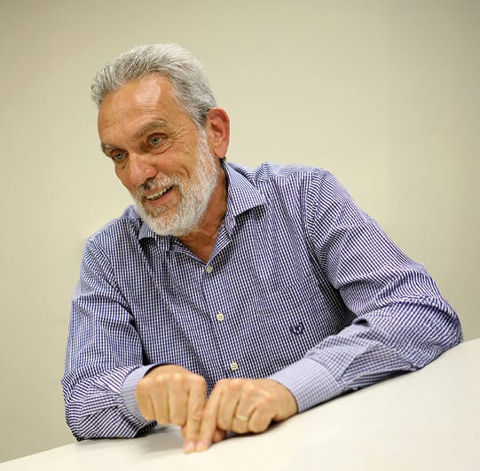Pessoa com cabelos grisalhos e barba, vestindo camisa de manga longa em tons de azul e branco com padrão xadrez, apoiada sobre uma mesa branca com ambas as mãos, em frente a fundo neutro bege. Pessoa com cabelos grisalhos e barba, vestindo camisa de manga longa em tons de azul e branco com padrão xadrez, apoiada sobre uma mesa branca com ambas as mãos, em frente a fundo neutro bege.