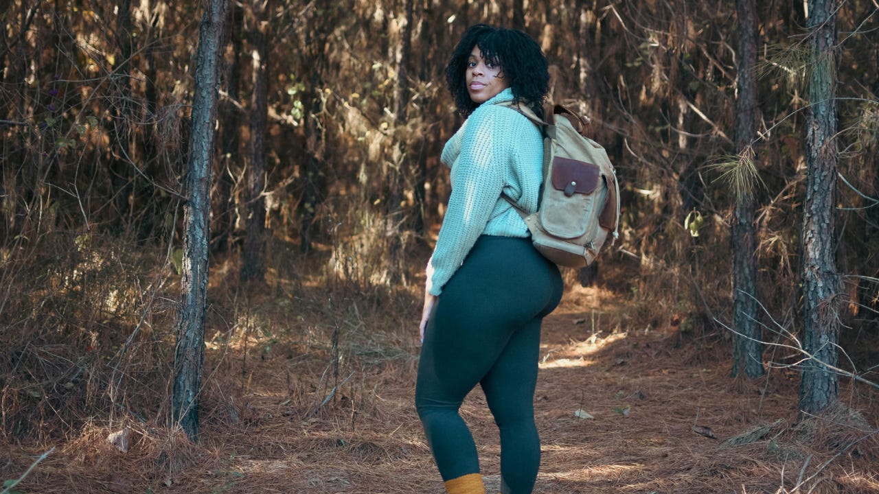 Plus-size person with a backpack standing on a pine-covered trail in a forest, looking back over their shoulder, wearing a light sweater, leggings, and boots, surrounded by tall trees and soft natural light. Plus-size person with a backpack standing on a pine-covered trail in a forest, looking back over their shoulder, wearing a light sweater, leggings, and boots, surrounded by tall trees and soft natural light.