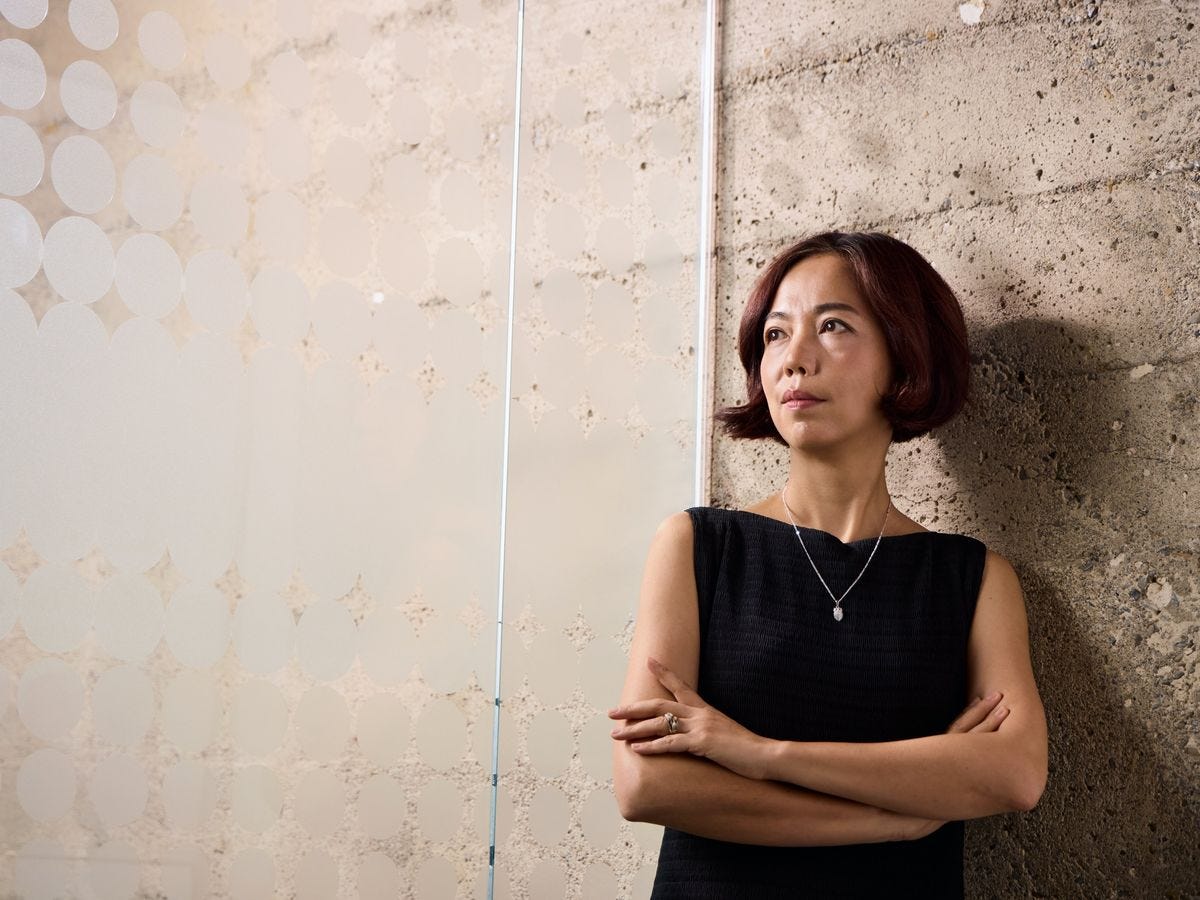 Fei-Fei Li wearing a black dress and posing against a concrete wall with arms crossed. Fei-Fei Li wearing a black dress and posing against a concrete wall with arms crossed.