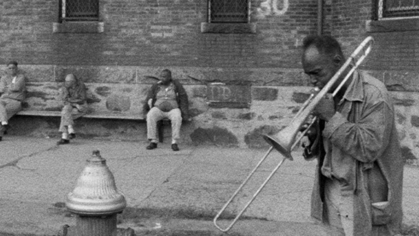 Titicut Follies | Still features patients sitting outside of a hospital, with one holding a trombone.