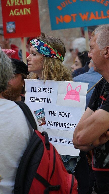 What Donald Trump Understands about American Men
Image description: a photo of a person with long curly hair wearing a colorful headband and holding a sign that reads "I'll be post-feminist in the post-patriarchy."
