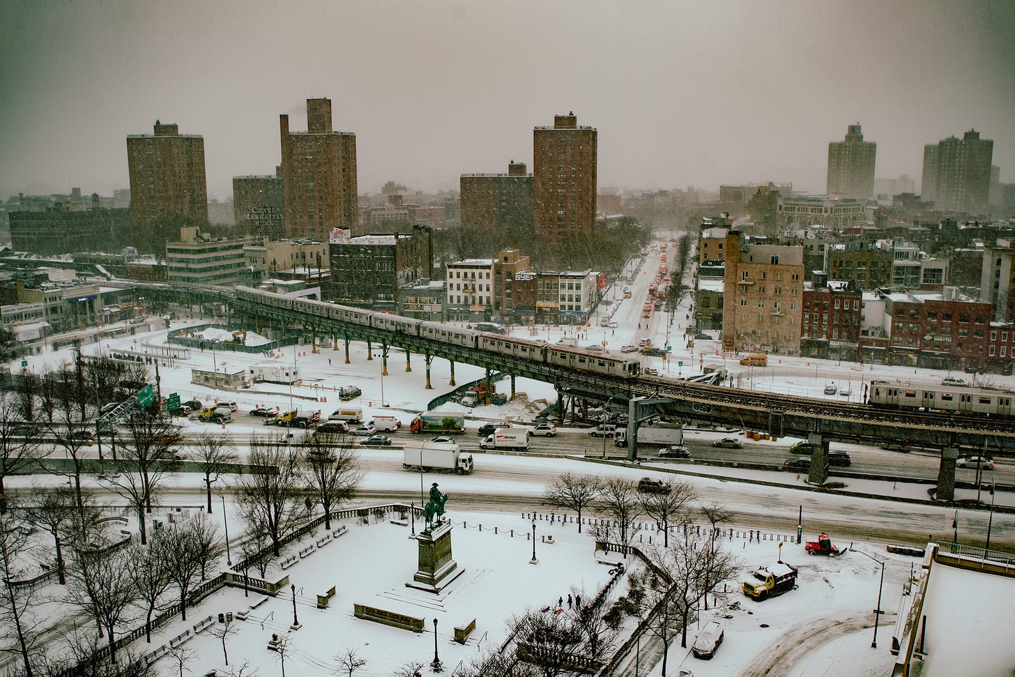 A wide, elevated view of a city neighborhood during a heavy snowstorm. An elevated subway train crosses through the middle of the frame, and below it, a park with a monument is covered in snow. The streets and buildings are all blanketed in white under a grey, overcast sky.