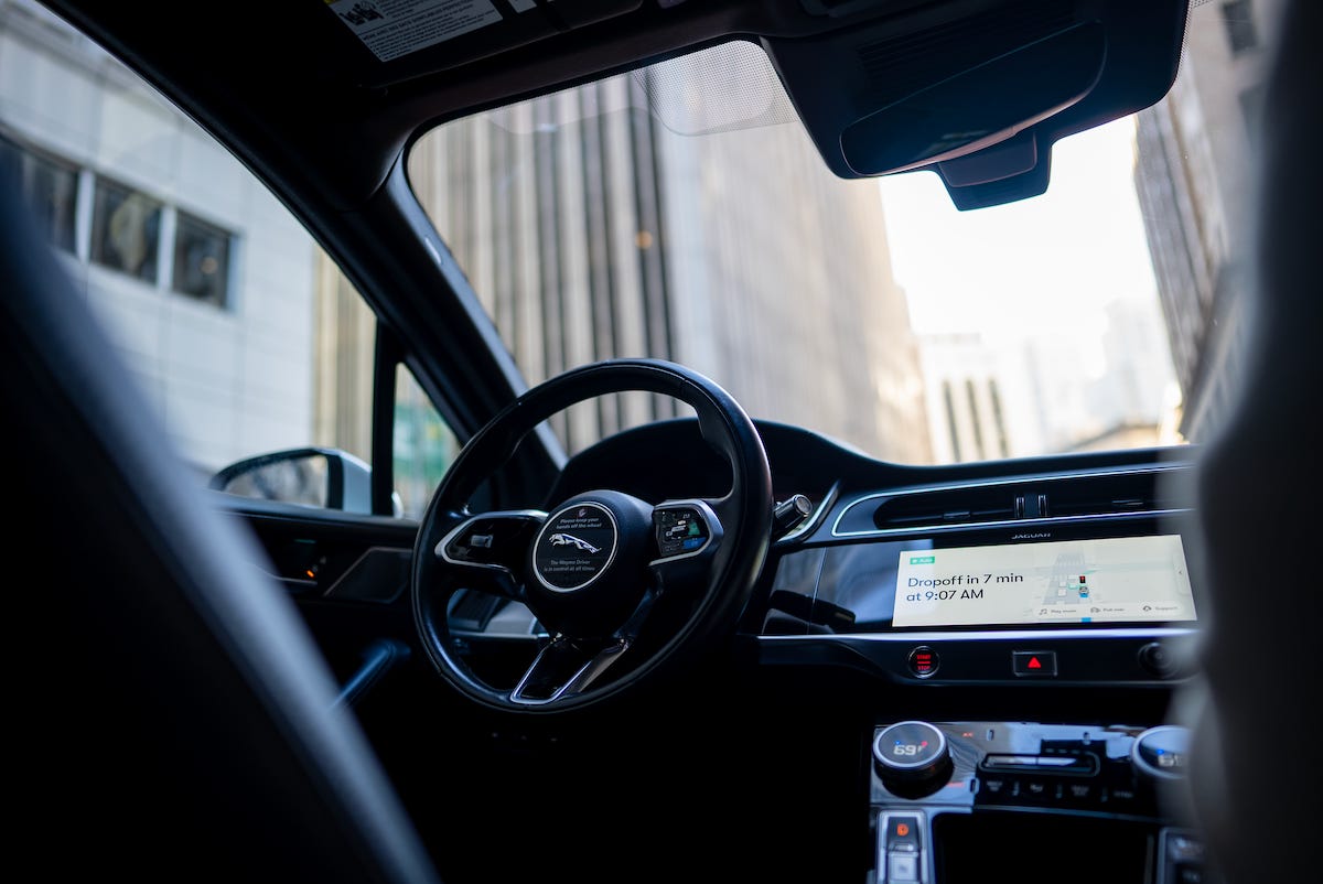 Interior view of a luxury vehicle showing a black steering wheel, digital dashboard display with navigation information showing a 7-minute dropoff time at 9:07 AM, and a bright urban street visible through the windshield. Interior view of a luxury vehicle showing a black steering wheel, digital dashboard display with navigation information showing a 7-minute dropoff time at 9:07 AM, and a bright urban street visible through the windshield.