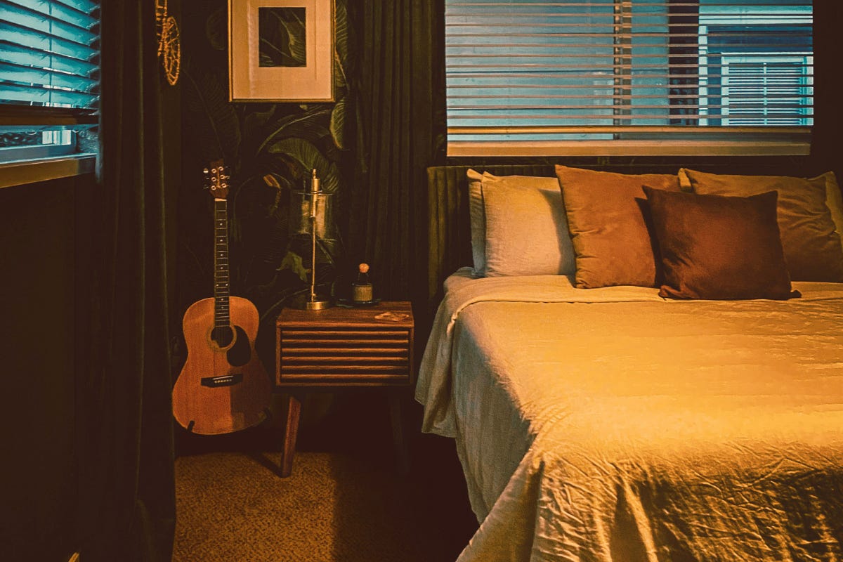 A cozy, dark-walled bedroom with warm lighting, featuring a bed with textured pillows, an acoustic guitar, and a mid-century modern nightstand. A cozy, dark-walled bedroom with warm lighting, featuring a bed with textured pillows, an acoustic guitar, and a mid-century modern nightstand.