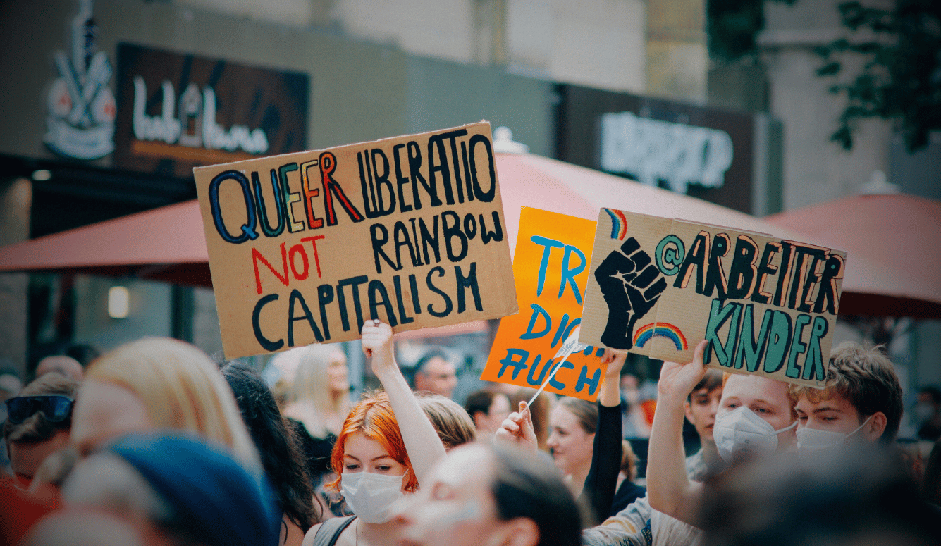 Protesters in face masks hold placards reading Queer Liberation Not Rainbow Capitalism and Arbeiter Kinder. Protesters in face masks hold placards reading Queer Liberation Not Rainbow Capitalism and Arbeiter Kinder.