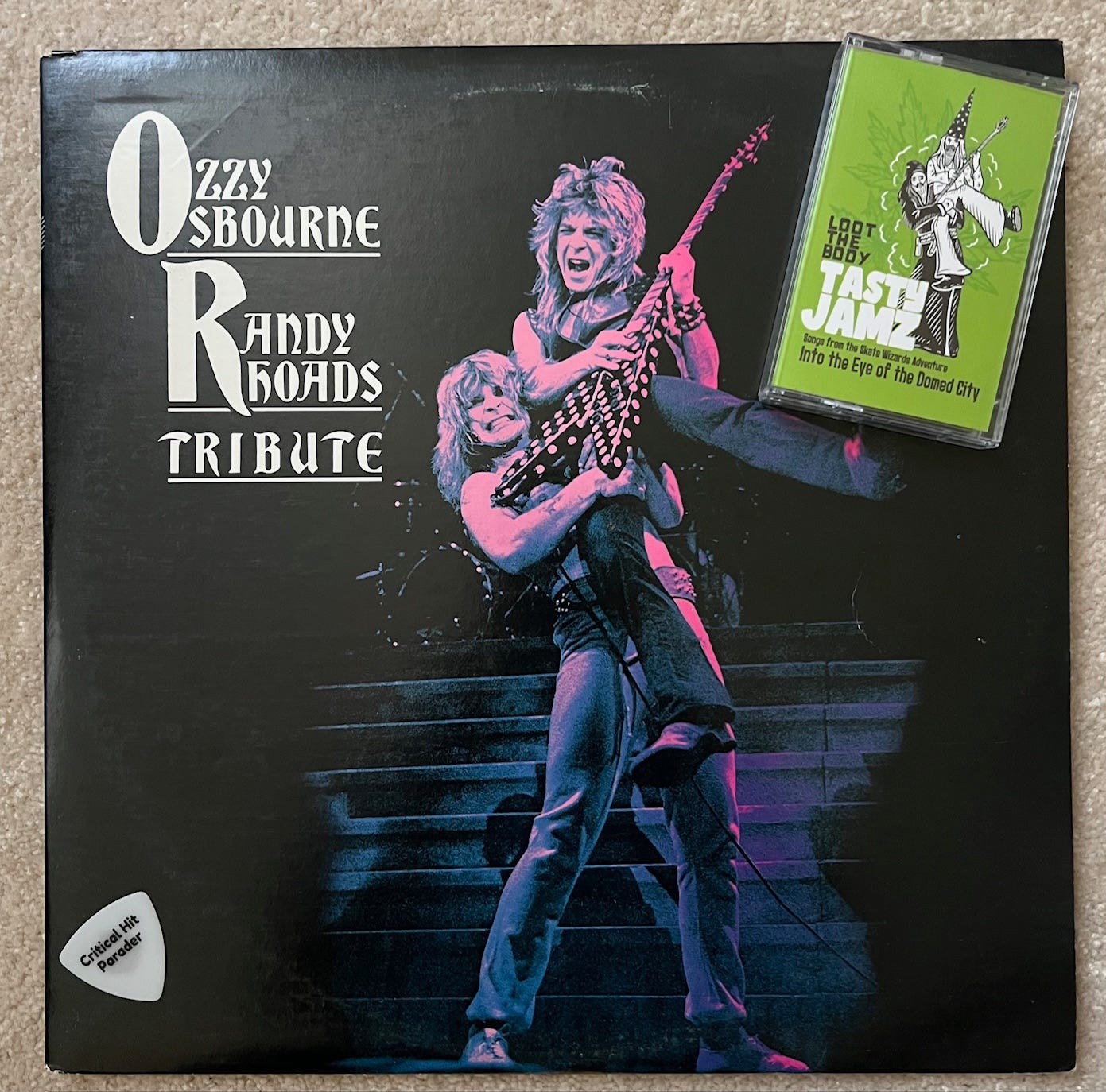 A Loot the Body Tasty Jamz cassette sits on the top right corner of the Ozzy Osbourne - Randy Rhoads Tribute vinyl album. The cassette features a black robed wizard holding up a white robed wizard playing a staff like a guitar on a bright green background. The album features Ozzy holding Randy in the exact same pose. A white Critical Hit Parader guitar pick sits reverently in tribute to the clever homage in the bottom left corner.