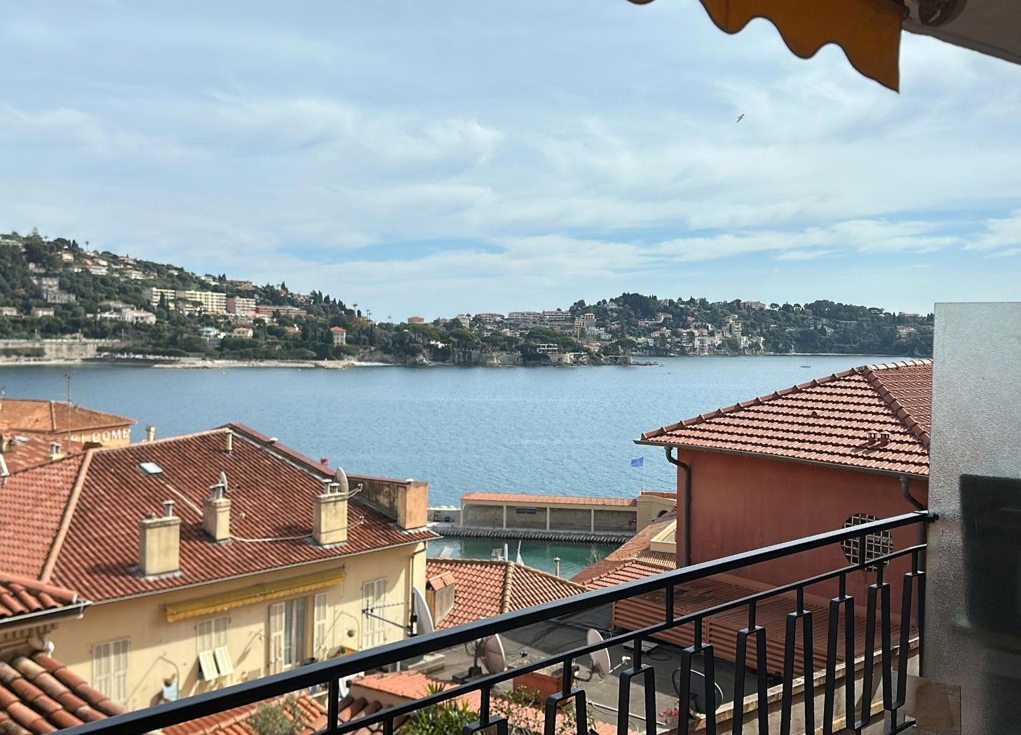 View from a balcony in Villefranche-sur-Mer: red tile roofs, a slice of the bay, hills across the water, an orange awning at the top of the frame. View from a balcony in Villefranche-sur-Mer: red tile roofs, a slice of the bay, hills across the water, an orange awning at the top of the frame.