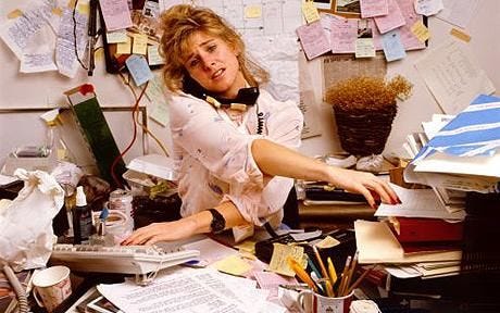 This may contain: a woman sitting at a desk with lots of clutter and papers on the wall
