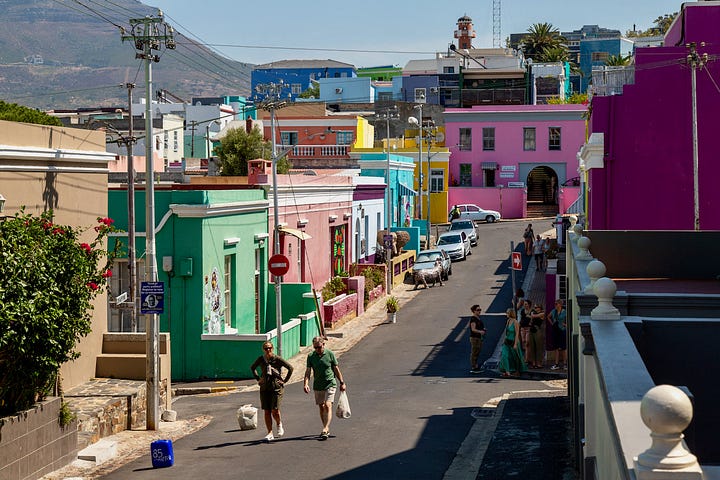 Four photographs from South Africa. Visitors stand on the hand-drawn floor map of old District Six inside the District Six Museum in Cape Town. A steep street of brightly painted houses in Bo-Kaap, with two people walking uphill carrying shopping. A Rovos Rail train alongside a platform at Capital Park station in Pretoria, with green and white branded umbrellas in the foreground. The interior of Clarke's Bookshop in Cape Town, seen from an upstairs landing, with crowded shelves and customers browsing tables of books.