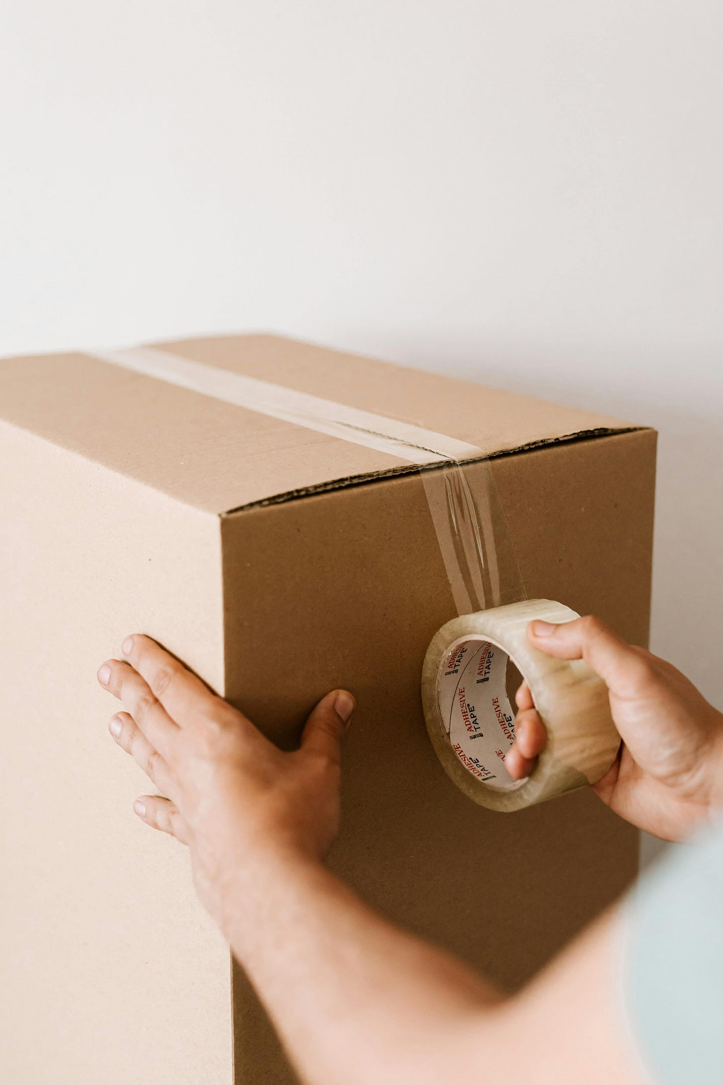 Two hands are shown placing packing tape on an unmarked cardboard box