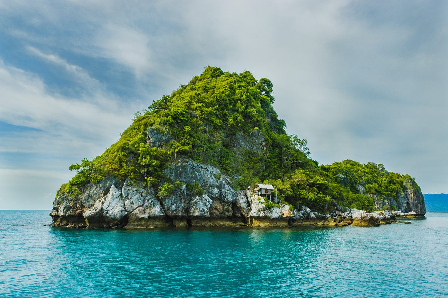 A rocky outcrop sits as a small island in clear blue water. A small hut sits along the shore of the mountain, nestled beside the rock face. A rocky outcrop sits as a small island in clear blue water. A small hut sits along the shore of the mountain, nestled beside the rock face.