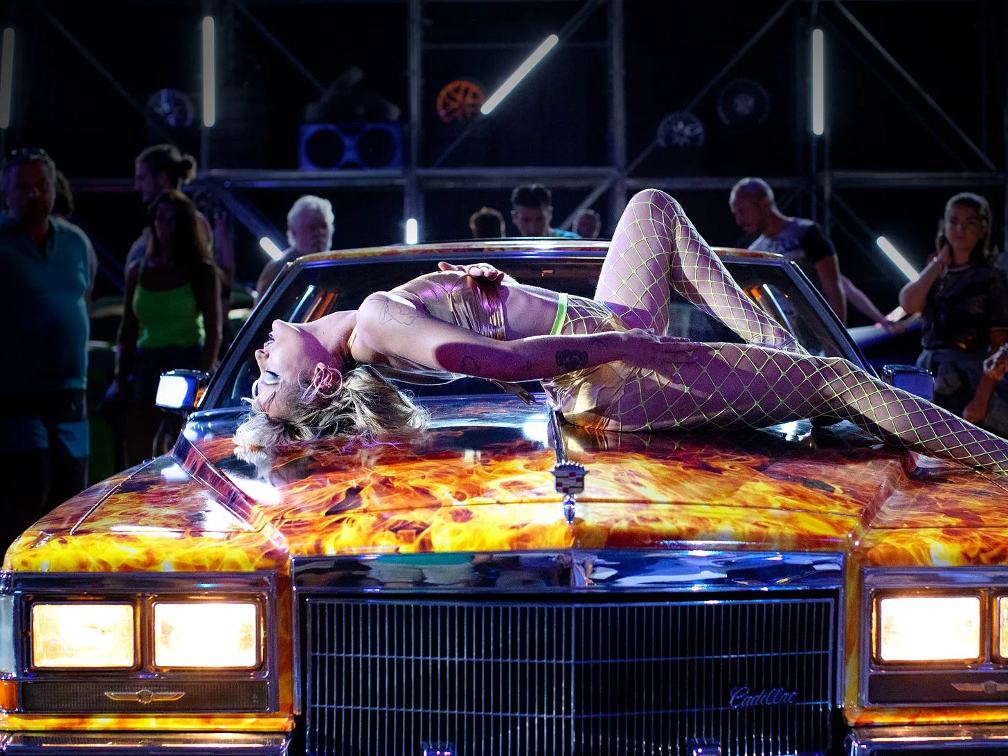scantily clad woman lying/writhing on the top of a car with flames painted on it