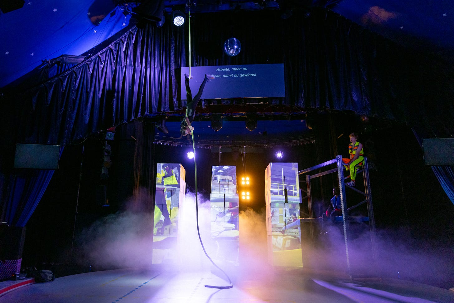 A performer hangs upside down from a rope from the ceiling of  a circus tent.