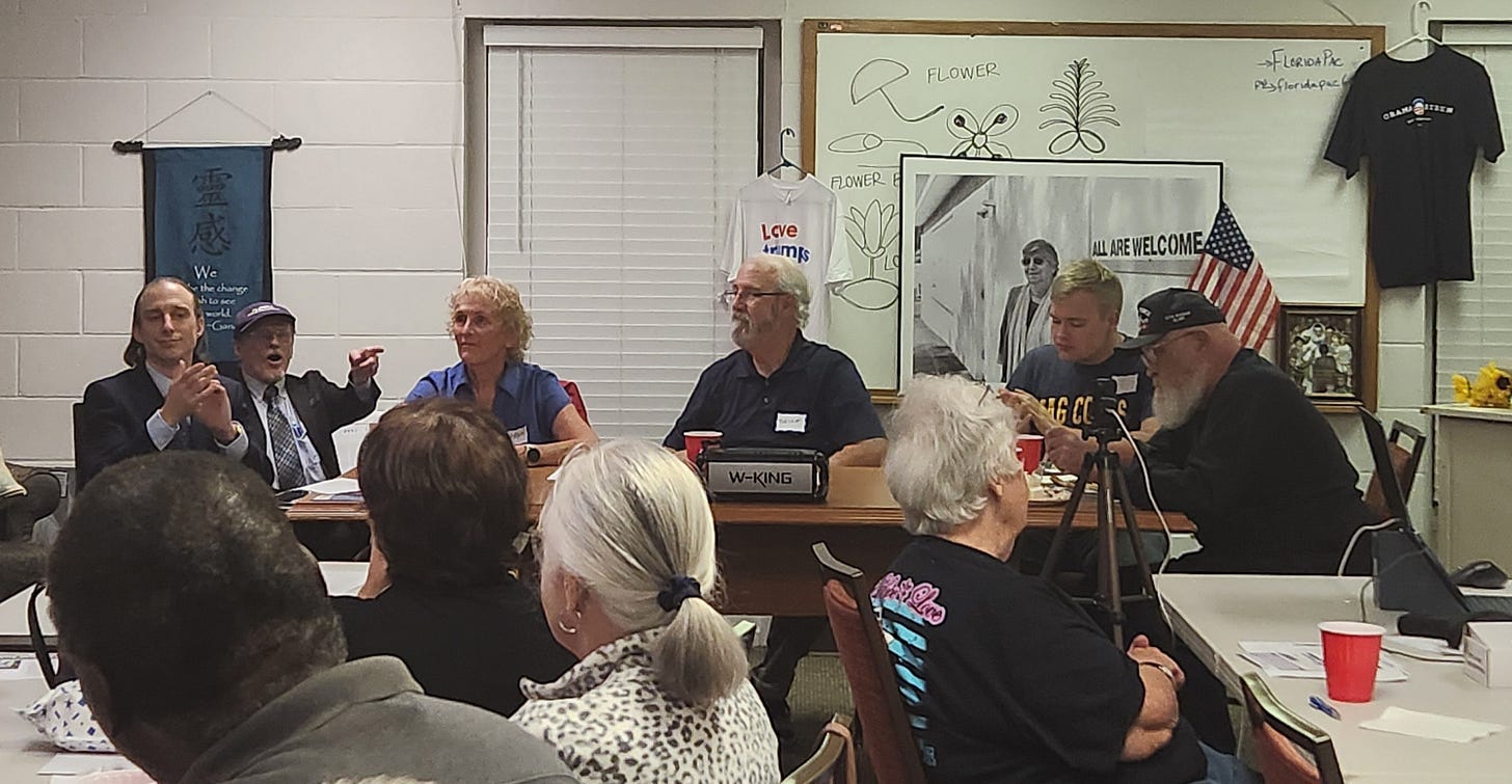 A community meeting or panel discussion in a small room. Five people sit at a front table facing an audience, with papers, red plastic cups, and a camera on a tripod recording. A black “W-KING” speaker sits on the table. On the walls are a blue banner with a quote, a whiteboard with flower sketches and the note “FloridaPac,” a grayscale poster reading “ALL ARE WELCOME,” a small U.S. flag, and two hanging T‑shirts: one says “Love trumps,” the other says “Obama/Biden.” Blinds cover the windows, and attendees of various ages watch and listen.
