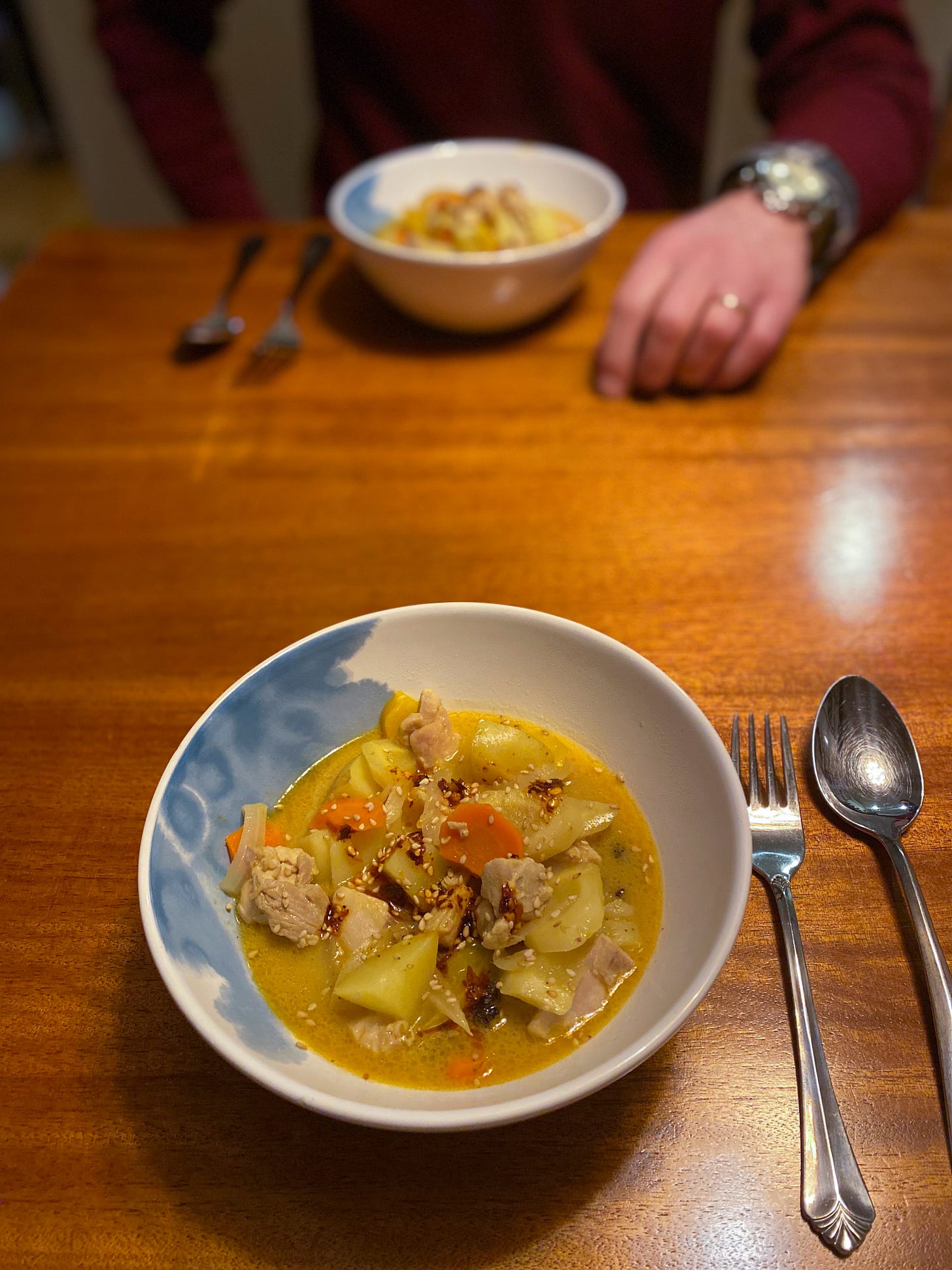 Two white and blue bowls of yellow curry with chicken, potatoes, carrots, & peppers across from each other on the table. The curry is topped with sesame seeds and chili oil, and next to each bowl are a fork and spoon. Jeff's hand rests next to his bowl across the table.