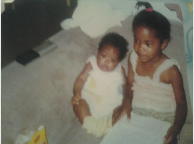Picture of a Black toddler and infant sitting on a sofa. The toddler has a notebook in her lap.