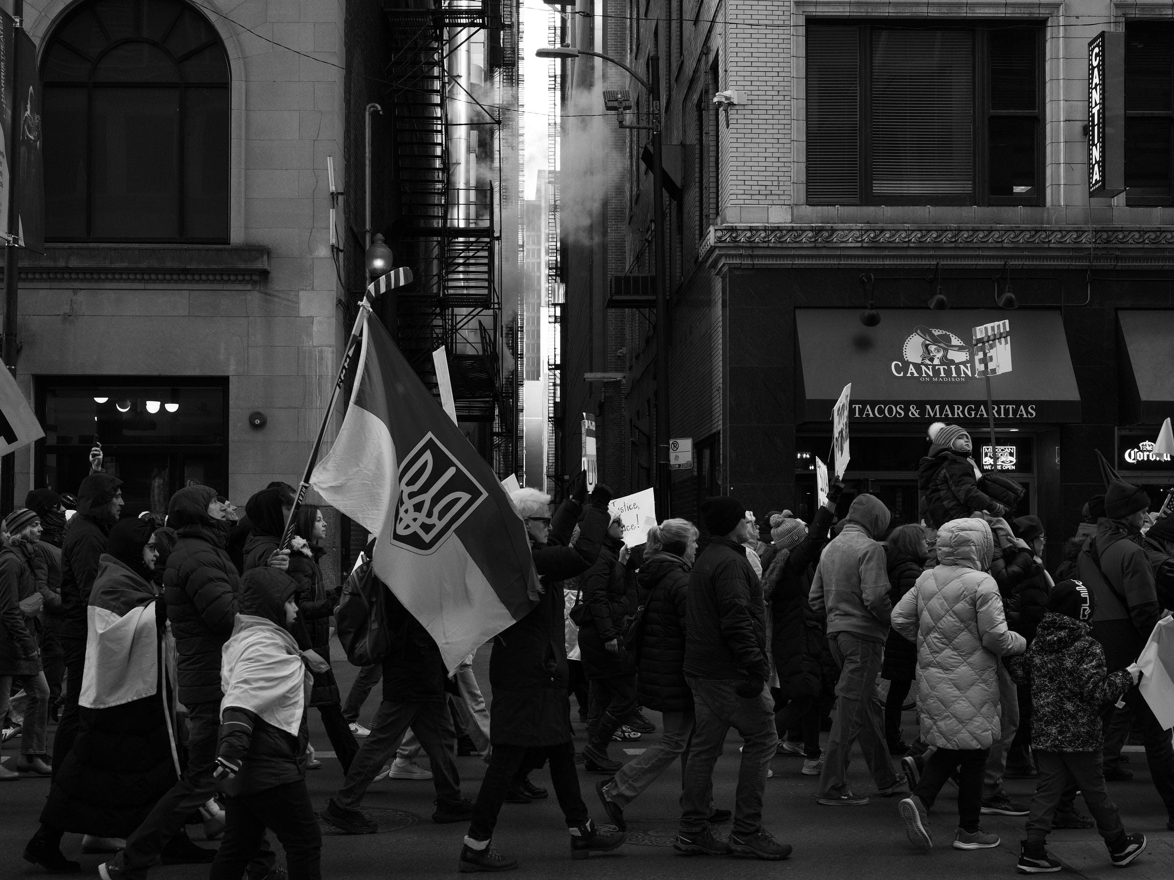 A black and white photograph captures a protest march with demonstrators carrying Ukrainian flags and signs through a city street. A large Ukrainian flag with a trident emblem is prominently displayed. Steam rises between tall buildings in the background, adding depth to the urban setting. Protesters, bundled in winter clothing, walk with determination, some carrying children on their shoulders. The storefront of a Mexican restaurant, "Cantina," is visible, contrasting with the serious tone of the demonstration. A black and white photograph captures a protest march with demonstrators carrying Ukrainian flags and signs through a city street. A large Ukrainian flag with a trident emblem is prominently displayed. Steam rises between tall buildings in the background, adding depth to the urban setting. Protesters, bundled in winter clothing, walk with determination, some carrying children on their shoulders. The storefront of a Mexican restaurant, "Cantina," is visible, contrasting with the serious tone of the demonstration.
