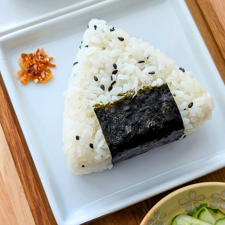 Single mushroom pâté-filled onigiri with crispy onion salt next to a pickled cherry blossom onigiri, highlighting two distinct Japanese rice ball styles.