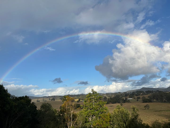 two photos of a rainbow: the first is looking through some trees as it comes up from the ground and the second is a long shot of a full rainbow above some low hills with a blue sky with some grey rain clouds above