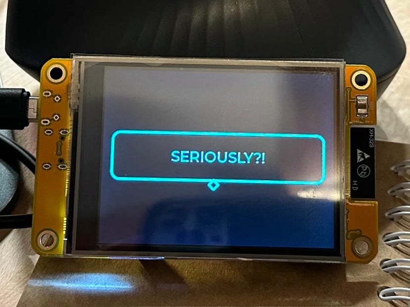 Photo of a yellow ESP32 touchscreen showing a glowing blue speech bubble with the word “SERIOUSLY?!” on a dark background.
