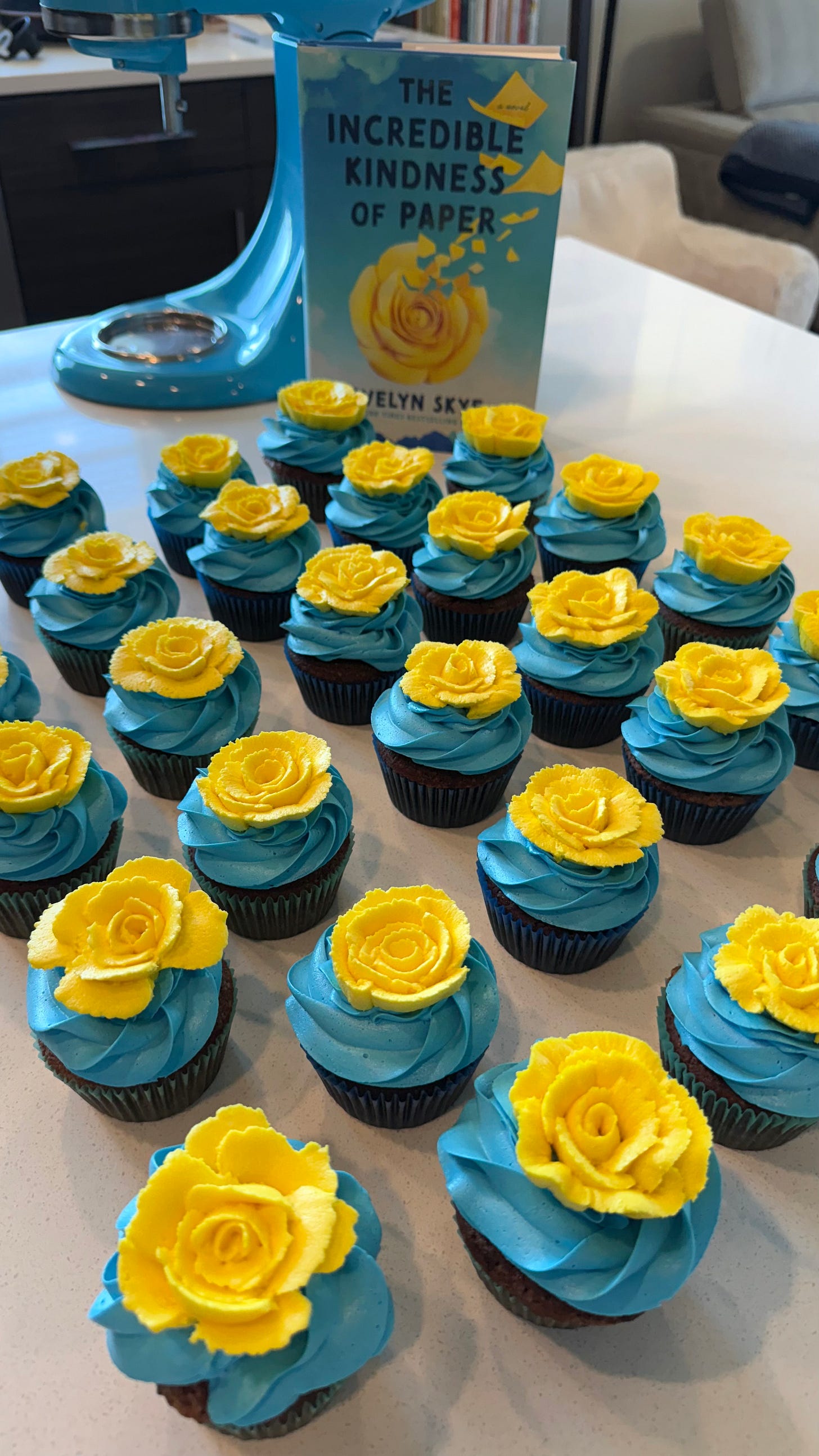 chocolate cupcakes with blue frosting and topped with yellow buttercream roses that match the book cover of The Incredible Kindness of Paper, in the background next to a blue mixer