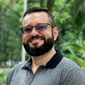 Homem de cabelo e barba pretas. Usa óculos, uma camisa polo cinza escuro e esboça um sorriso. Homem de cabelo e barba pretas. Usa óculos, uma camisa polo cinza escuro e esboça um sorriso.