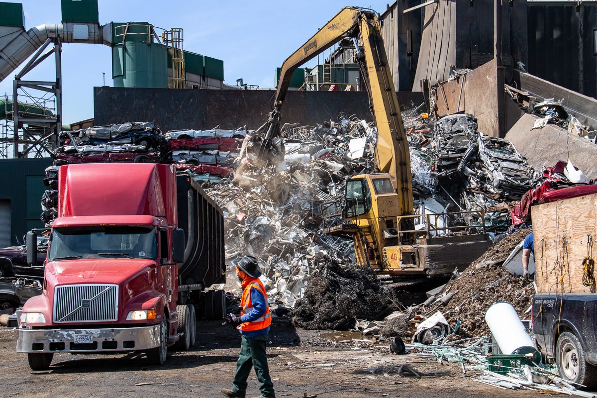 Yellow excavator with claw attachment unloading metal scrap into a pile near a red semi-truck and worker in orange safety vest.