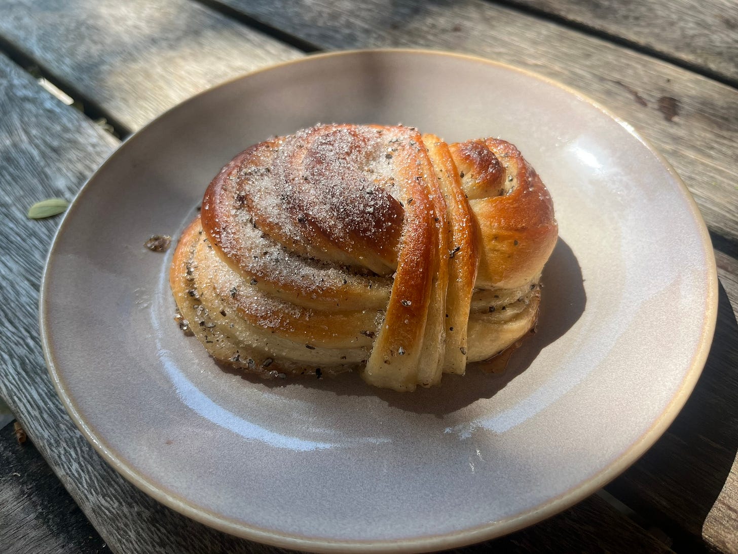 A knotted cardamom bun with powdered sugar and crushed cardamom on top, on a beige, round plate on a wooden table