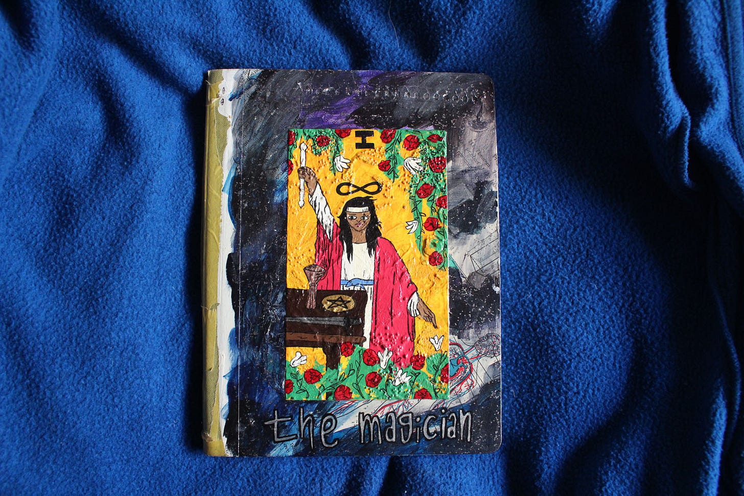 a dark, painted composition book. it's purple with a drawing of the magician tarot card