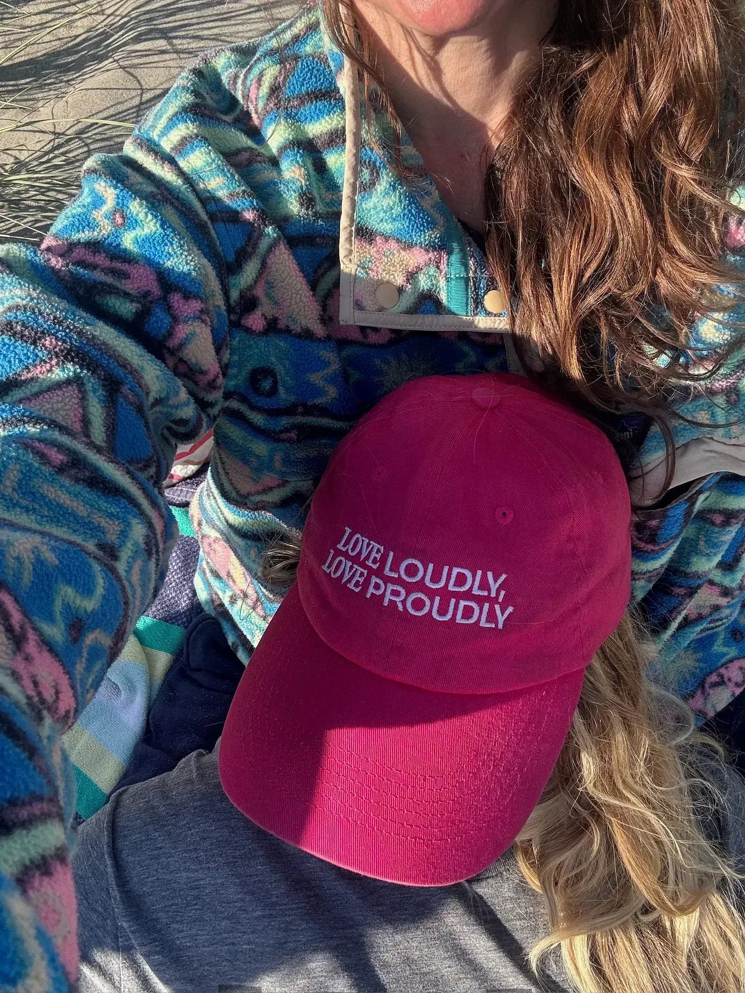 a person lies on another person’s lap, wearing a bright pink hat reading “LOVE LOUDLY, LOVE PROUDLY,” over long blond hair.